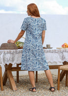 Woman stands backwards wearing a 100% cotton dress in white based blue floral print with short sleeves and one hand in pocket