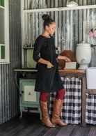 Woman standing side on in a black cotton corduroy above knee dress with hand in pocket, paired with maroon leggings and tan boots