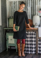 Woman in a black cotton corduroy above knee dress standing in a rustic kitchen holding a book.