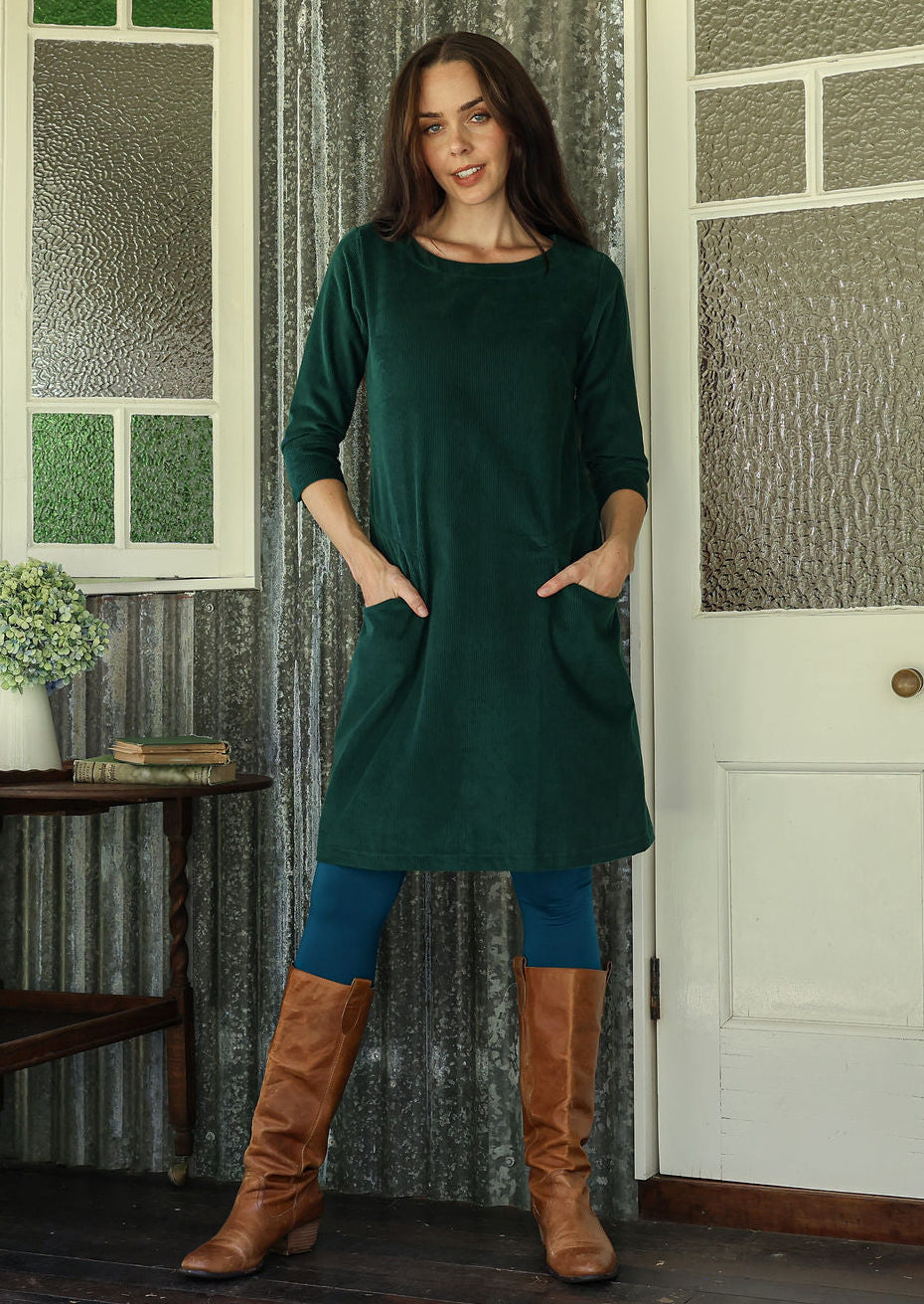 Woman wearing green corduroy A-line dress with 3/4 sleeves with hands in pockets over leggings and boots