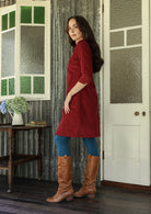 Woman standing sideways wearing a red corduroy dress and brown knee-high boots standing in a room with a door and window.