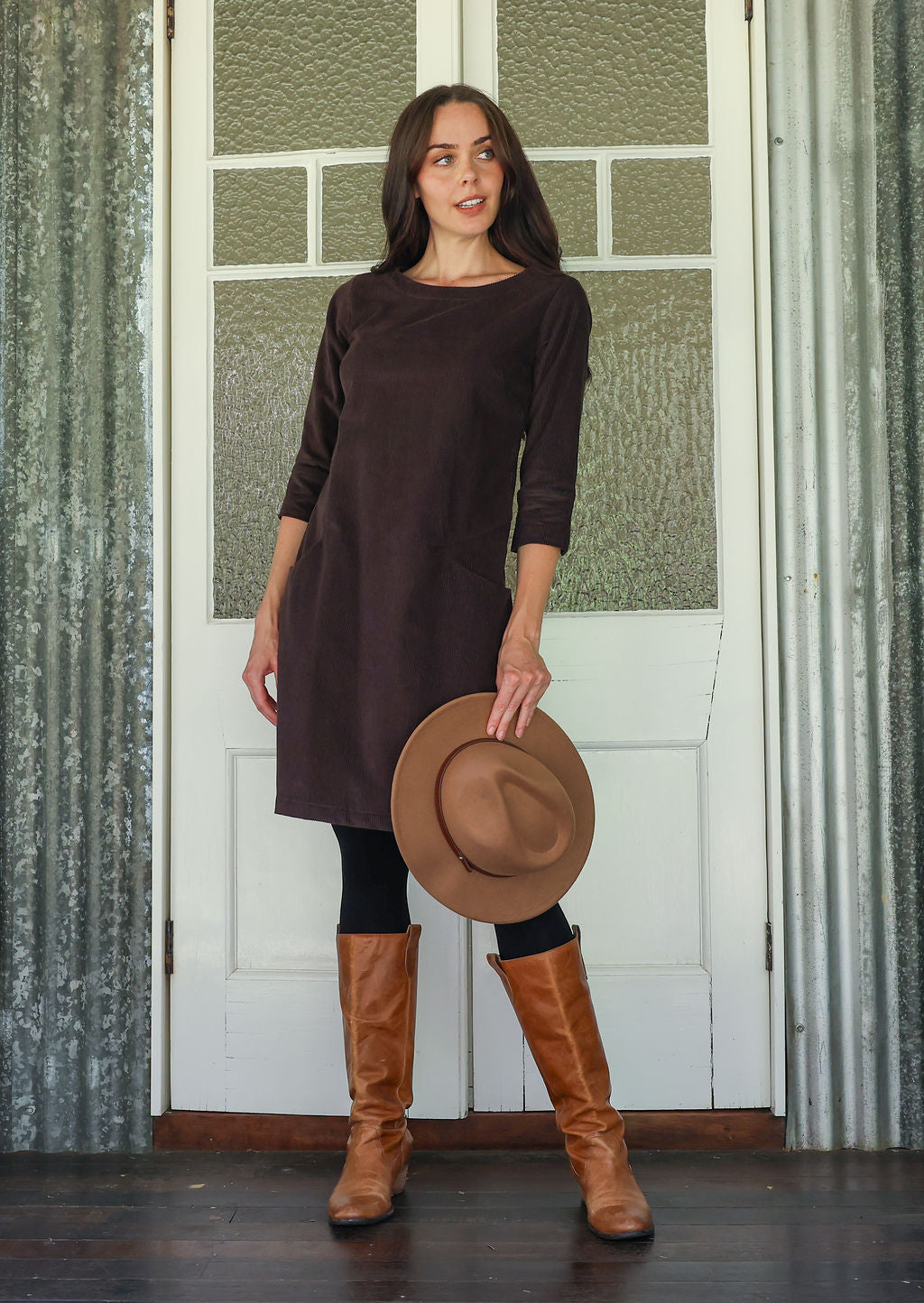 Woman wearing a brown corduroy dress and boots holding a hat in front of a white door with green window panes.