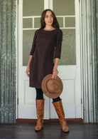 Woman wearing a brown corduroy dress and boots holding a hat in front of a white door with green window panes.