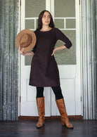 Woman in a brown dress and boots holding a hat against a textured wall.