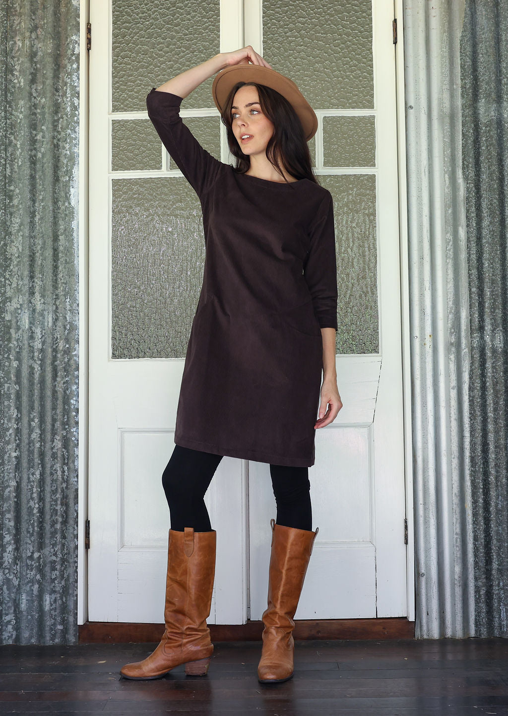 Woman wearing a dark brown dress, brown boots, and a beige hat indoors.
