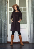 Woman wearing a dark brown A-line tunic dress 3/4 sleeves with hands in pockets  brown boots standing in front of a white door.