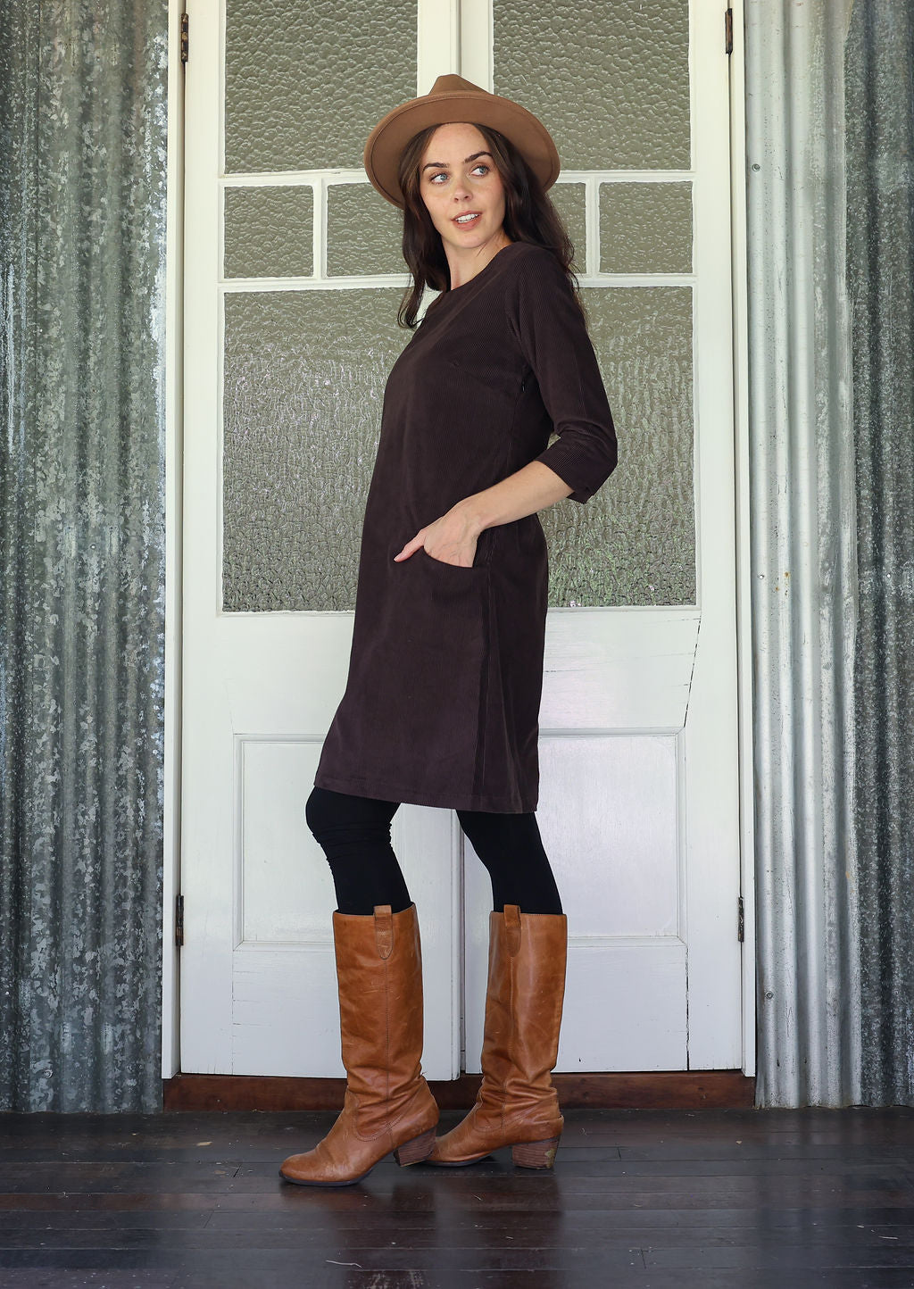 Woman standing sideways wearing a brown dress, hat, and boots standing in front of a white door with glass panels.