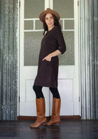 Woman standing sideways wearing a brown dress, hat, and boots standing in front of a white door with glass panels.