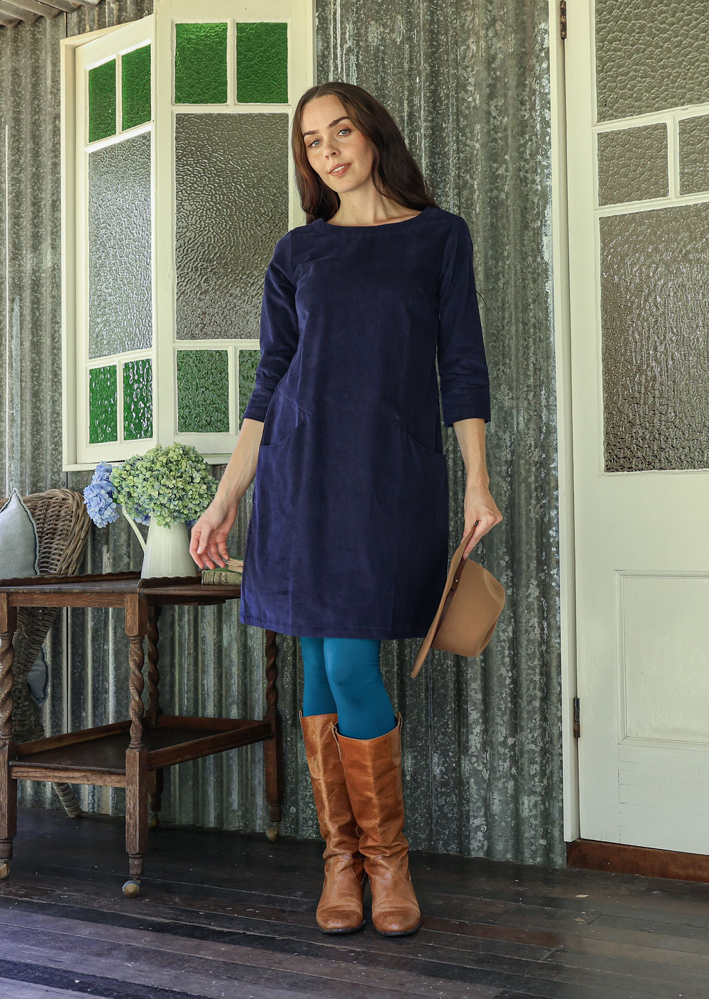 Woman in a navy blue corduroy dress and brown boots standing in a room with patterned wallpaper.
