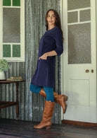 Woman standing sideways wearing a blue corduroy dress and brown boots standing in a room with patterned curtains and a door.