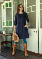 Woman in a navy blue corduroy dress with brown boots and a hat standing in a doorway.