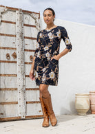 Woman wearing a black based floral print dress with brown boots standing against a rustic wooden door.