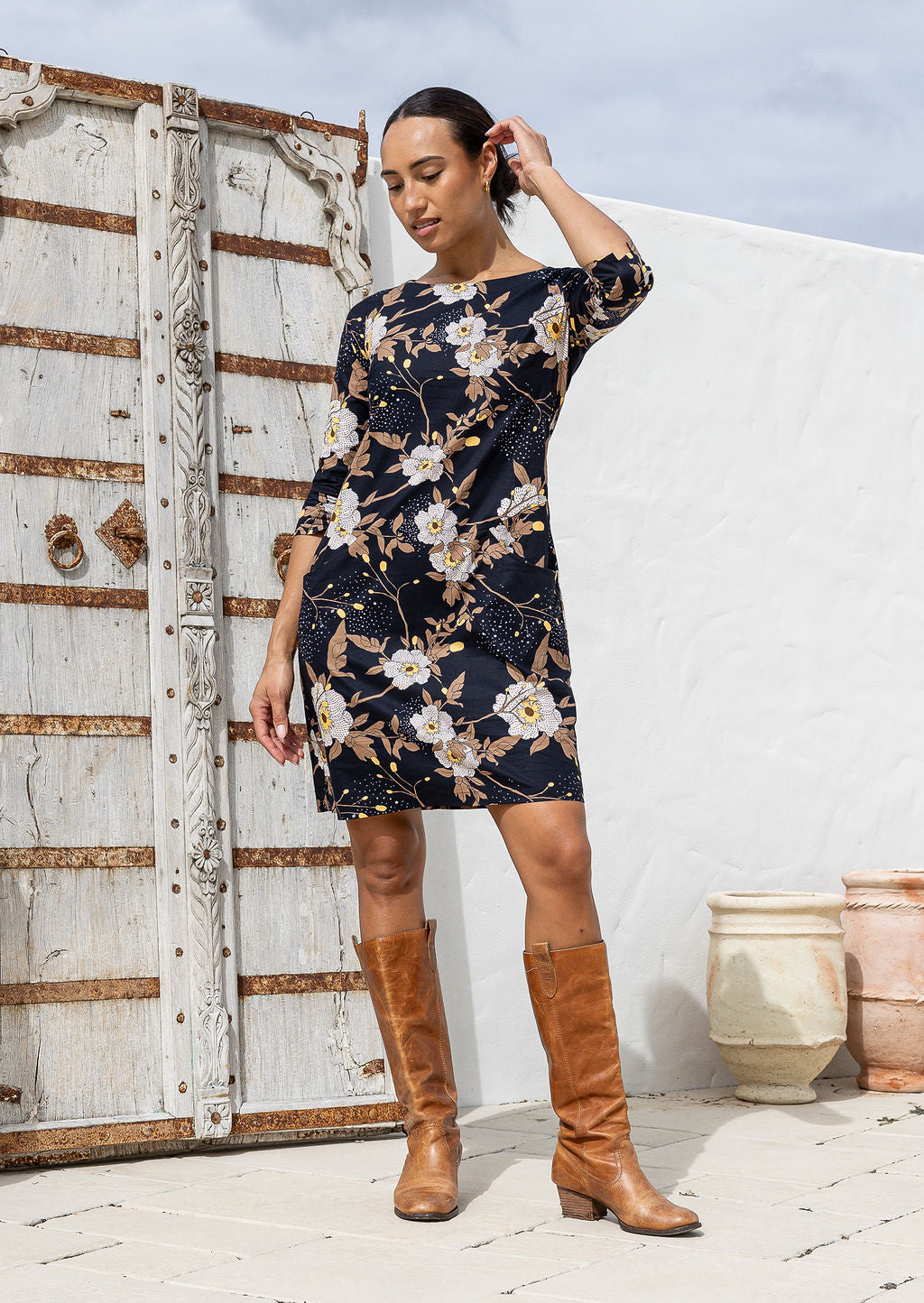 Woman wearing a floral dress and brown boots standing against a white wall with a wooden door.
