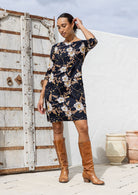 Woman wearing a floral dress and brown boots standing against a white wall with a wooden door.