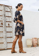 Woman standing sideways wearing a floral dress and brown boots standing against a white wall with decorative elements.