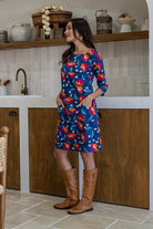 Woman standing sideways wearing a floral dress and brown boots standing in a kitchen setting.