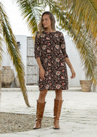 Woman wearing a black based floral print dress with brown boots standing against a rustic wooden door.