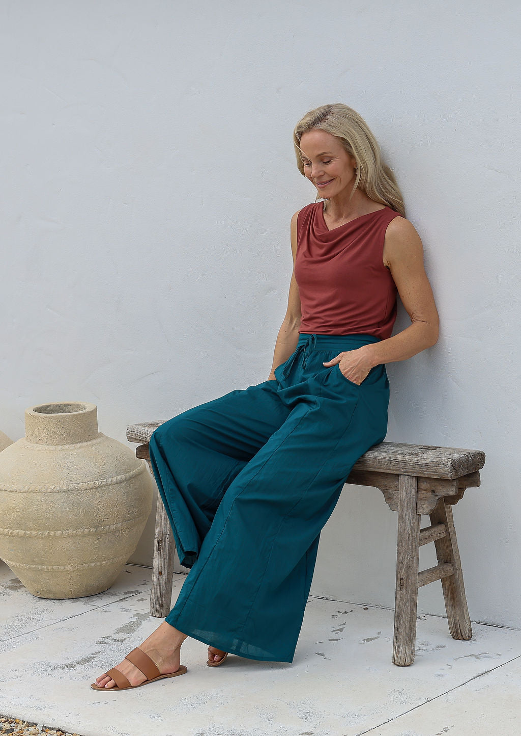 Woman sitting on a wooden stool wears 100% cotton wide leg pants in deep teal with pockets, elastic waist and drawstring tie, paired with terracotta basic top.