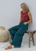 Woman sitting on a wooden stool wears 100% cotton wide leg pants in deep teal with pockets, elastic waist and drawstring tie, paired with terracotta basic top.