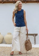 Woman wearing a blue sleeveless top and wide legged white cotton pants, holding a woven bag, standing against a white wall with decorative vases.