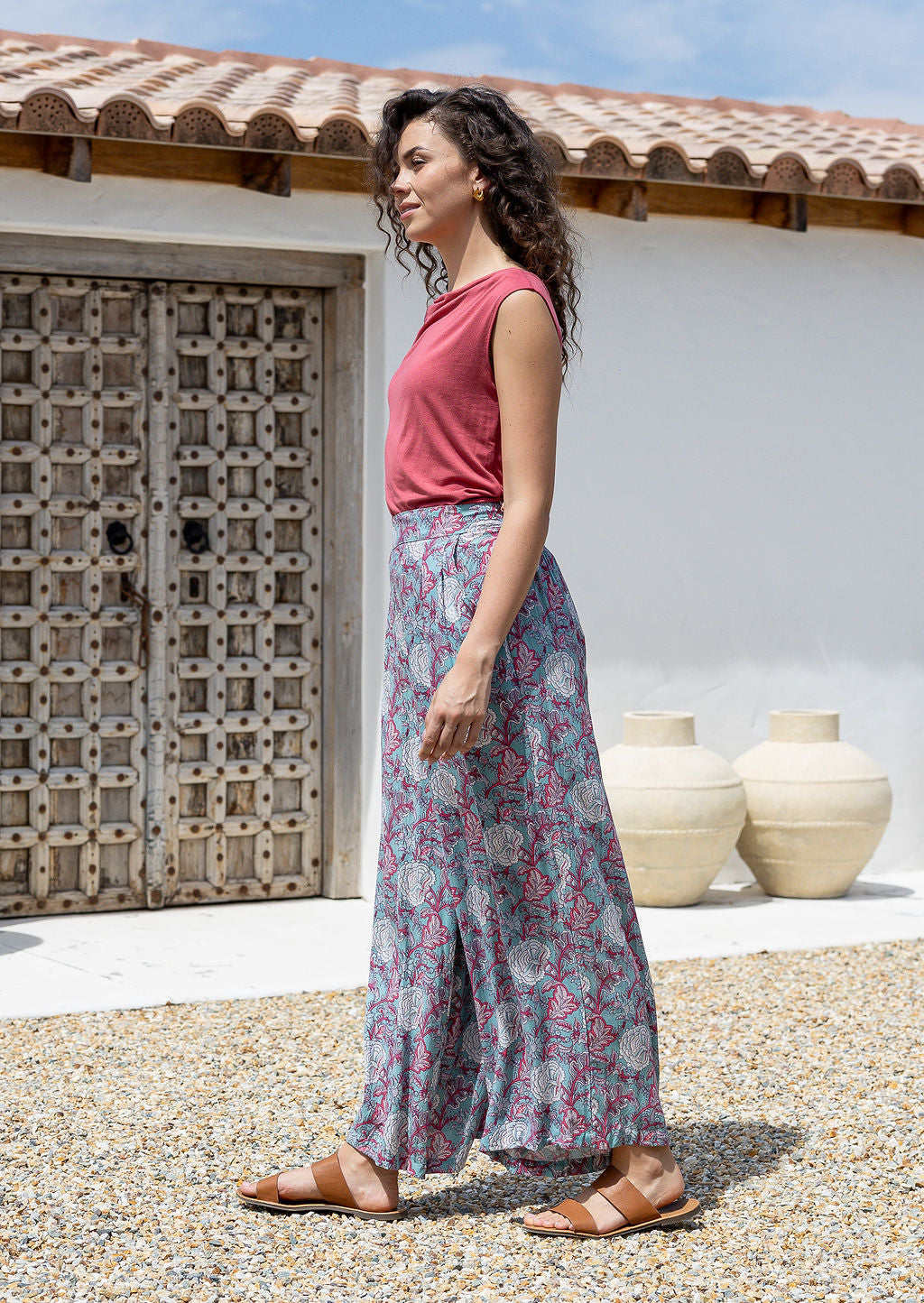 Woman wearing a pink top and rayon pants standing outdoors with a white wall and wooden door in the background.