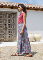 Woman wearing a pink top and rayon pants standing outdoors with a white wall and wooden door in the background.