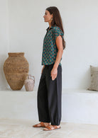 Woman stands sideways wearing a green based floral cotton top with short folded cuff sleeves, collar and V neckline 