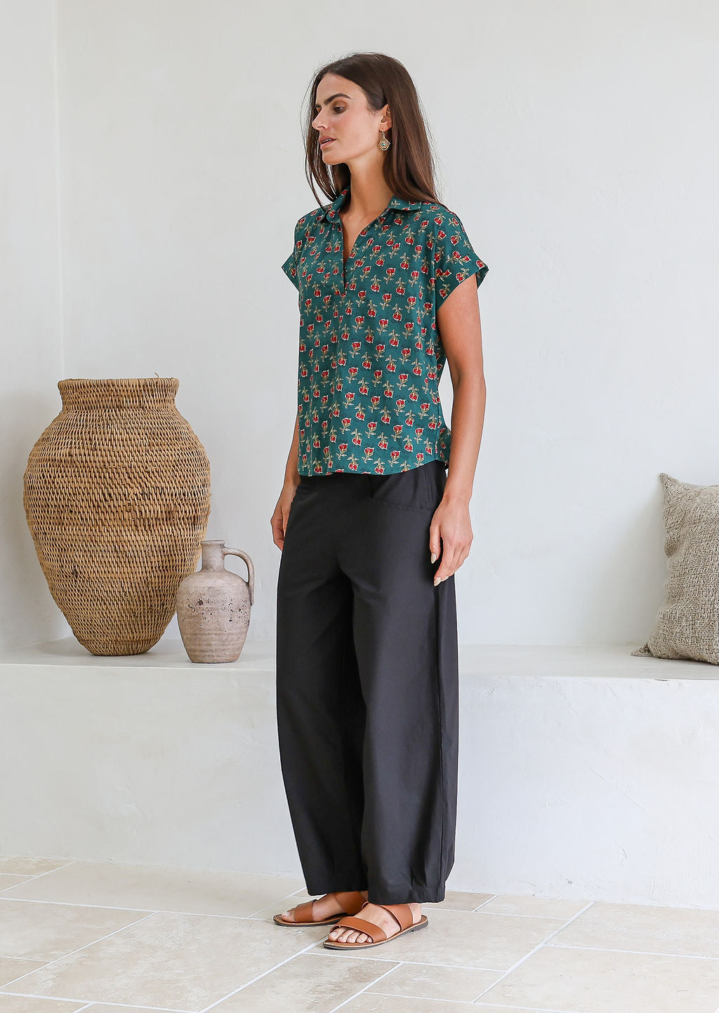 Woman wearing a bottle green based floral 100% cotton top and black pants standing in a room with decorative vases.