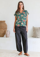 Woman wearing a green based floral print  100% cotton top and black pants in a minimal indoor setting.