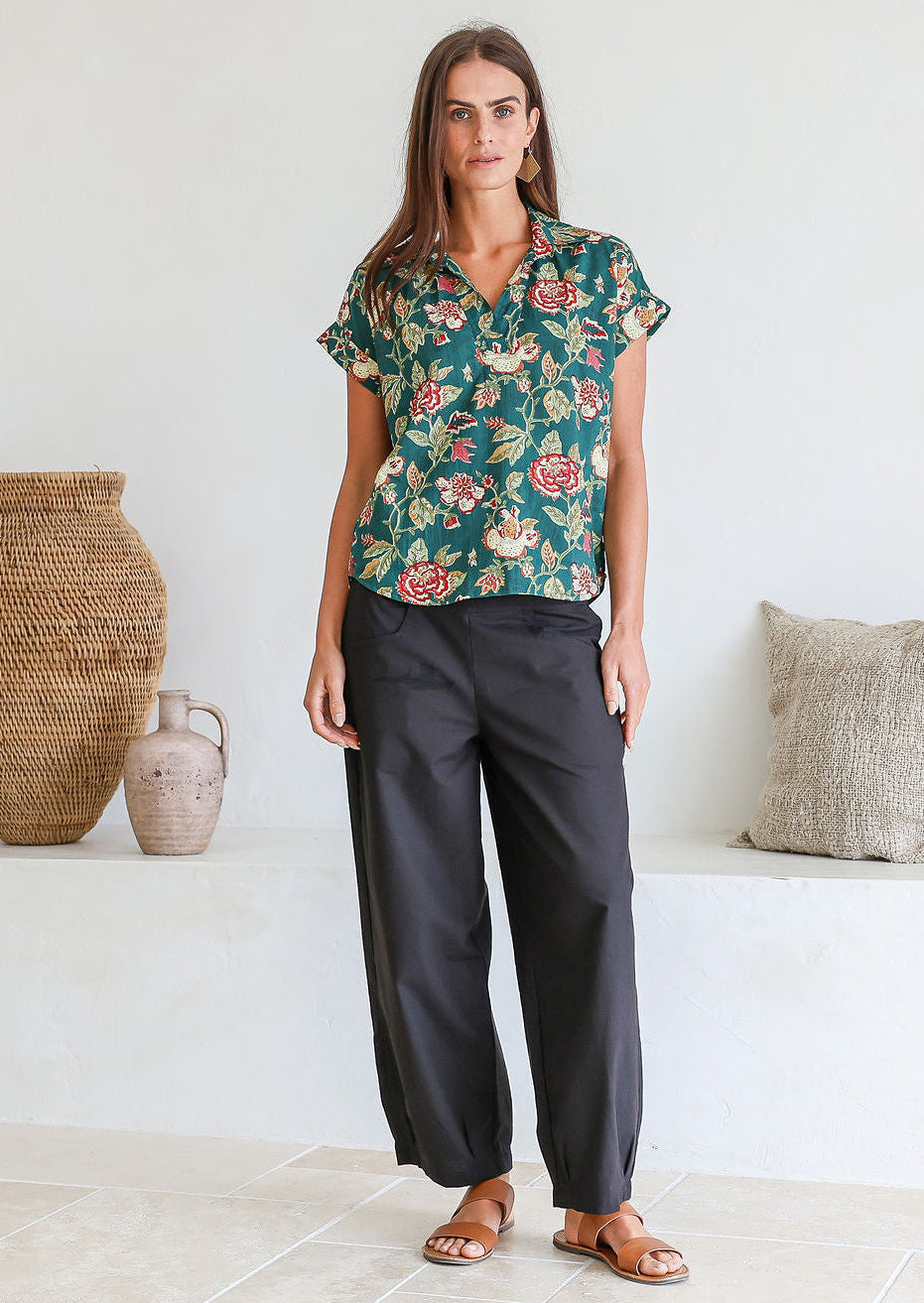 Woman wearing a green based floral print  100% cotton top and black pants in a minimal indoor setting.
