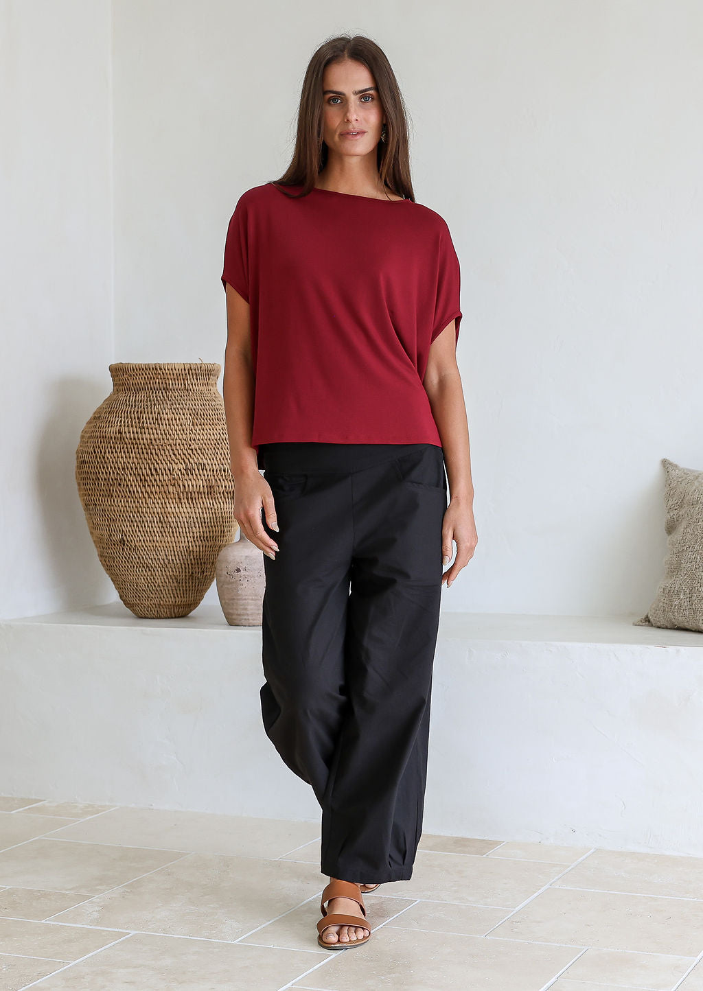 Woman wearing a maroon stretch rayon short sleeved batwing top and black pants standing in a room with decorative vases.
