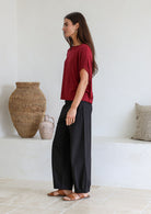 Woman standing sideways wearing a maroon relaxed fit stretch rayon t-shirt and black pants against a white wall.