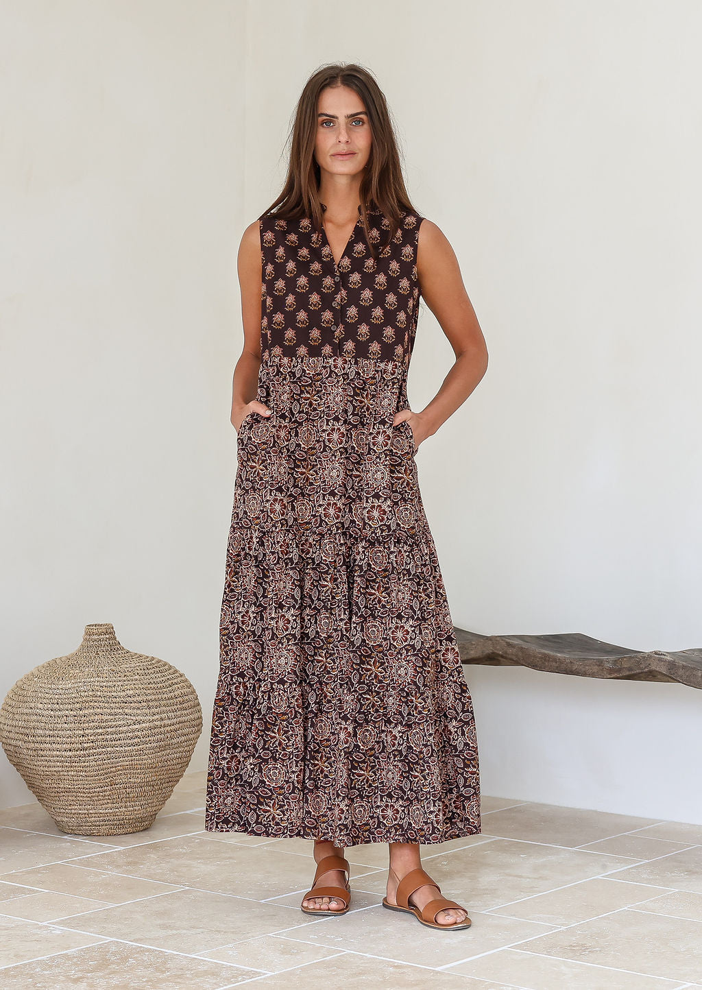 Woman wearing a Indian patterned cotton dress standing in a minimalistic room with a woven vase and wooden bench with her hands in her pockets 
