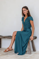 Woman in a teal rayon wrap dress sitting on a wooden stool against a white wall.