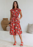 Woman wearing a midi length red based floral cotton dress with buttoned bodice, standing against a white wall and decorative vases