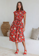 Model stands with wearing a midi length floral printed shirt style cotton dress with cap sleeves, buttoned bodice and a removable waist tie