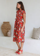 Model stands sideways near wearing a mid length, shirt style floral print cotton dress with blue, teal, green, pink, orange and tan on a on a red base.