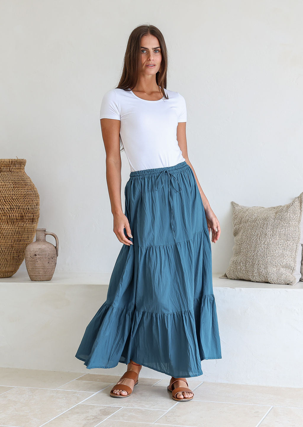 Woman wearing a white simple top and deep blue skirt with elasticated waistband standing in a minimalistic room.