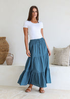 Woman wearing a white simple top and deep blue skirt with elasticated waistband standing in a minimalistic room.