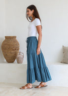 women stands sideways wearing a 100% cotton corsair blue skirt with a white plain top standing against a white background with decorative vases.