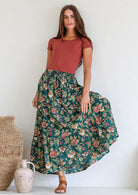 Woman wearing a 100% cotton floral maxi skirt and pink top standing against a white wall with decorative vases.