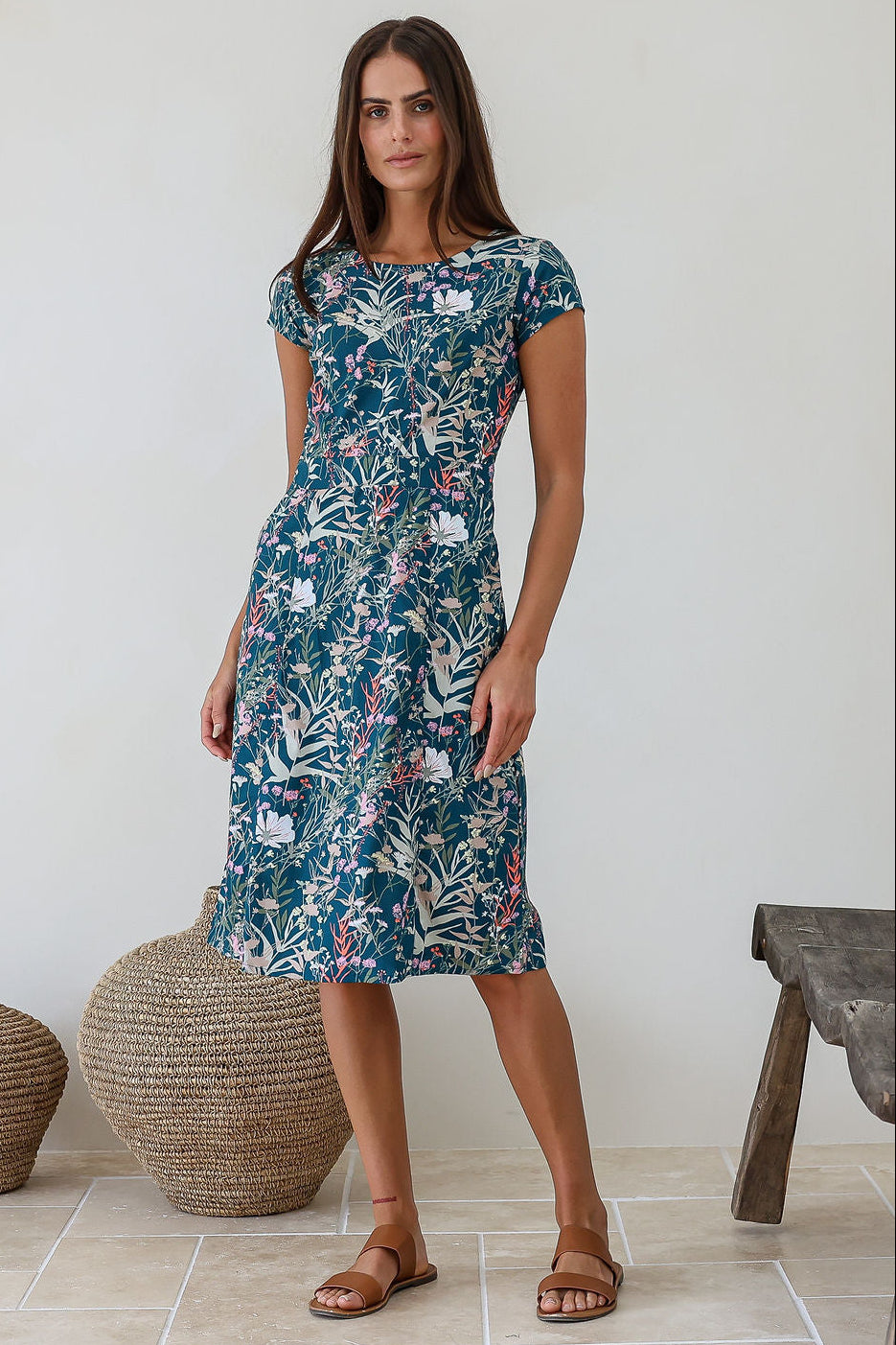Woman wearing a 100% cotton cap sleeved knee length dress with high round neckline, empire waist and slight a-line skirt in teal floral print.