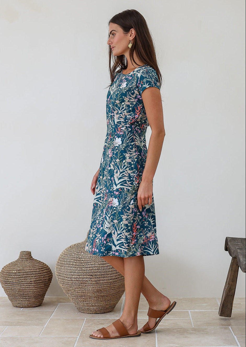 Woman stands sideways wearing a short sleeve sundress standing in a room with decorative vases and a wooden stool.