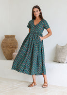 Woman standing with one hand in pocket wearing a relaxed fit midi length cotton dress with empire waistline and gathered design.