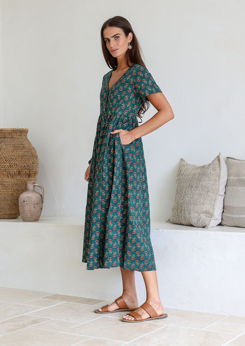 Woman stands sideways with one hand in pocket of a 100% cotton relaxed fit dress in bottle green with short sleeves, pockets and drawstring waist tie.