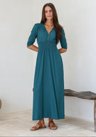 Woman wearing a teal rayon maxi dress with a buttoned bodice and elbow length sleeves, standing against a white wall.