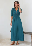 Woman wearing a teal rayon maxi dress standing in a minimalistic room with a wooden bench.
