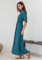 Woman standing side on wearing a teal rayon maxi dress with elbow length sleeves and a ruched waistband