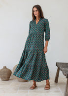 Woman standing against the white wall wears 3/4 sleeved cotton tiered maxi dress with mandarin collar and buttoned bodice 
