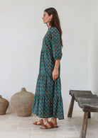Woman standing in front of wooden bench wears three tiered cotton maxi dress with 3/4 sleeves, pockets and mandarin collar